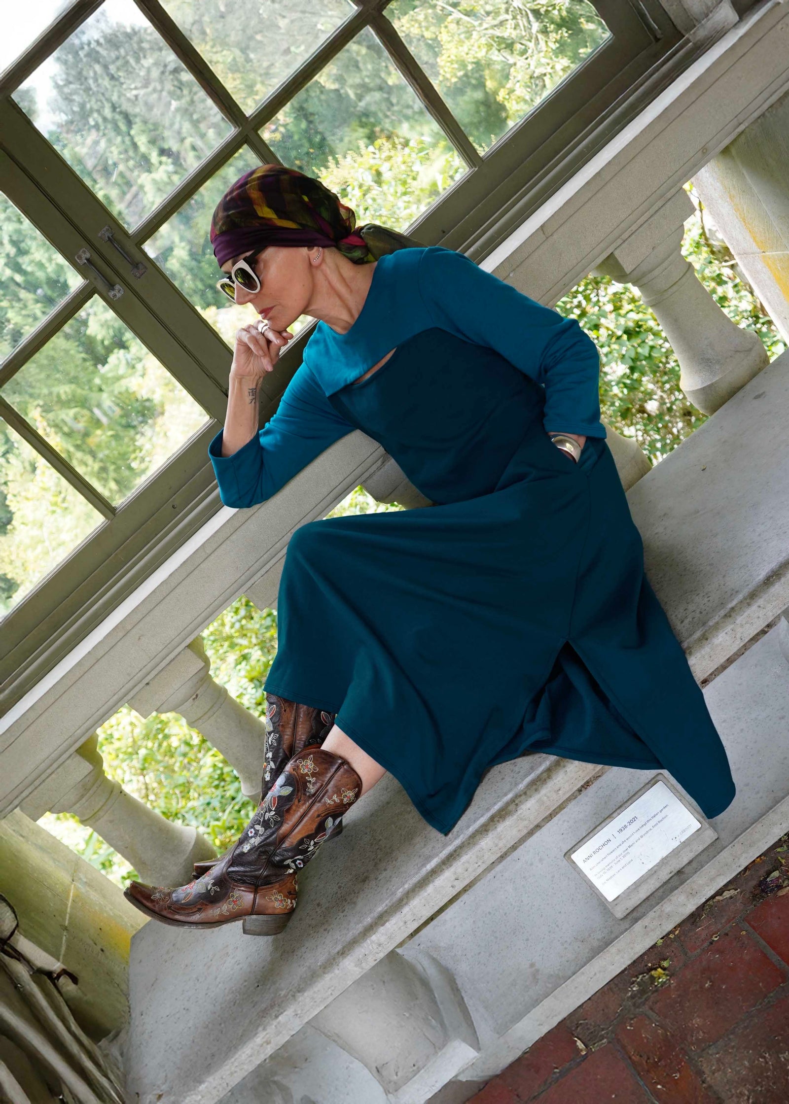 Woman in a blue dress and cowboy boots sitting on a stone ledge with greenery in the background