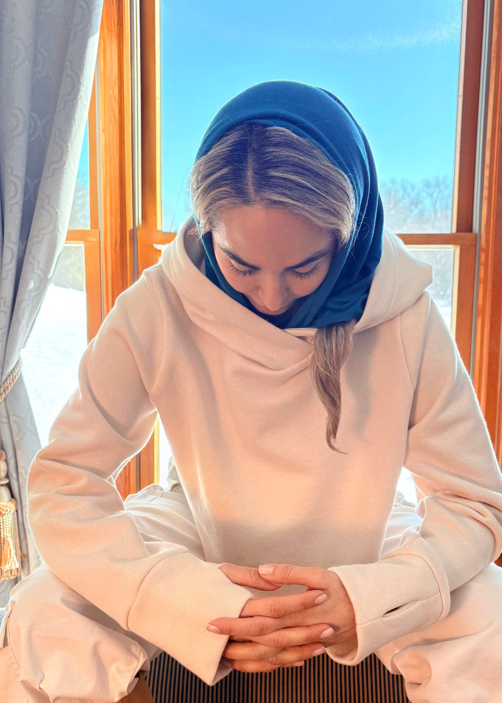 Person wearing a blue headscarf and beige outfit sitting by a window with a snowy landscape outside.