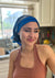 A beautiful lady in a jewel tone tank top, stands in her kitchen smiling. On her head she wears A paisley pattern of Deep blue with hints of golden, on a custom printed scarf, which is Made into a one piece headscarf with an organic cotton base in the sam